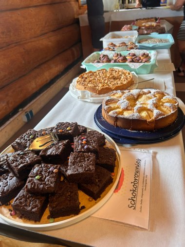 Auf einem langen Tisch mit weißer Tischdecke stehen verschiedenste Kuchen. Dahinter ist eine Holzwand.