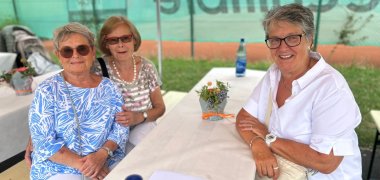 Jubiläum 30 Jahre Kita Hickelhäusje An einer Bierzeltgarnitur mit weißer Tischdekce sitzen drei Frauen mit Brille und Sonnenbrillen. Auf dme Tisch stehen Wasserflaschen und Blumendeko und im Hintergrund ist der grüne Banner am Zaun des Tennisplatzes.