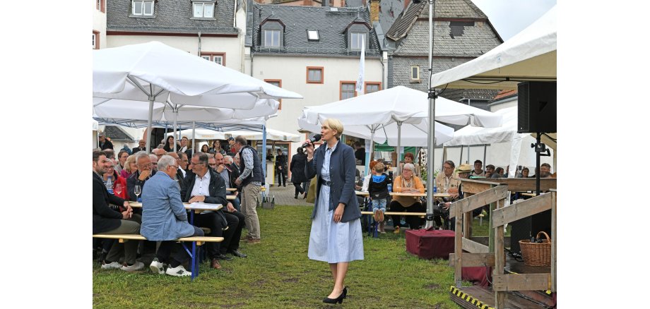 Auf einer Wiese stehen Bierzeltgarnituren und weiße, große, aufgespannte Sonnenschirme. Die Bänke sind besetzt und im Hintergrund ist das Rathaus und ein Weinstand zu sehen. Vorne steht eine Frau mit blonden Haaren, einnem hellbauen Kleid, einer blauen Jacke und blauen Pumps. Sie hält ein Mikrofon in der Hand und singt.