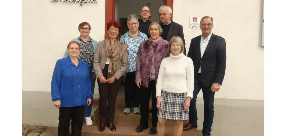 Dienstagsclub feiert sein 50-jähriges Bestehen Vor einer hellen Hauswand mit einer geöffneten Tür steht Bürgermeister Winfried Steinmacher, Diakon Hans-Jürgen Siebers und Pfarrer Ralph Senft und sechs Damen.