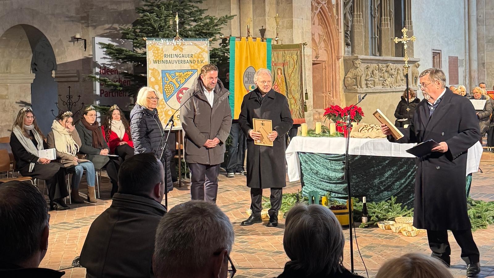 In einer Basilika stehen drei Männer und eine Frau vor einem Tisch, der mit einem grünem Samttuch und einer weißen Tischdecke gedeckt ist. Links davon sitzen vier Weinköniginnen in Winterjacken mit Kronen aauf dem Kopf. Daneben hängen drei große Stofffahnen vor einem großem Tannenbaum. Vorn im Bild sieht man Publikum, bestehend aus fünf HInterköpfen.