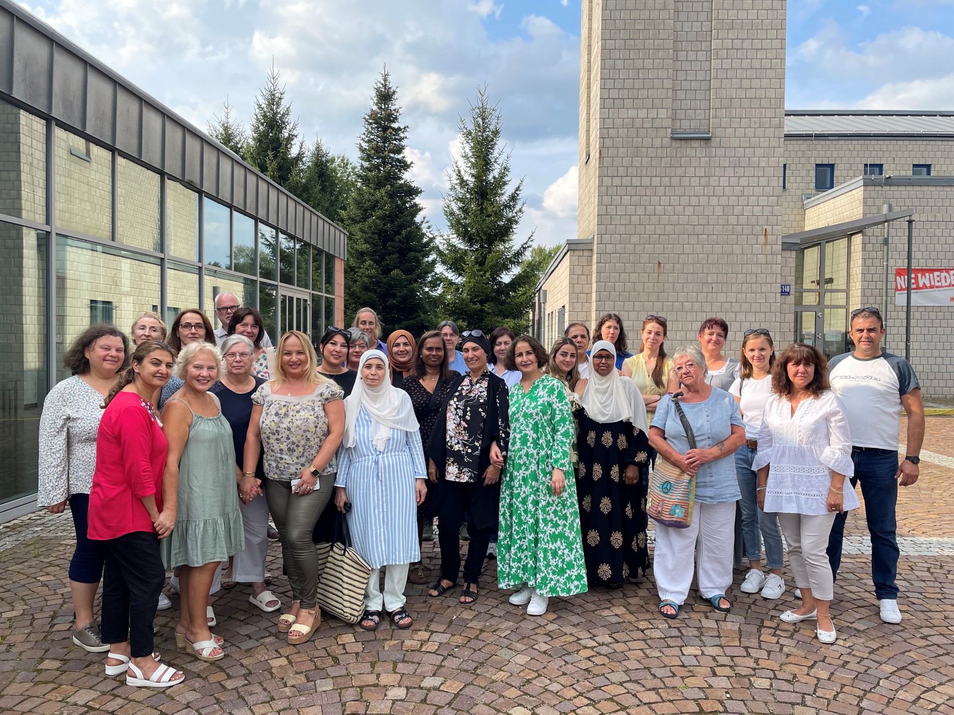 Eine Gruppe von 29 Personen stehen auf Kopfsteinpflaster vor einem aus hellen Steinen gemauertem Gebäude mit Turm. Links ist ein Gebäude mit großer Glasfront und im Hintergrund sind hohe Nadelbäume und ein bewölkter Himmel. 