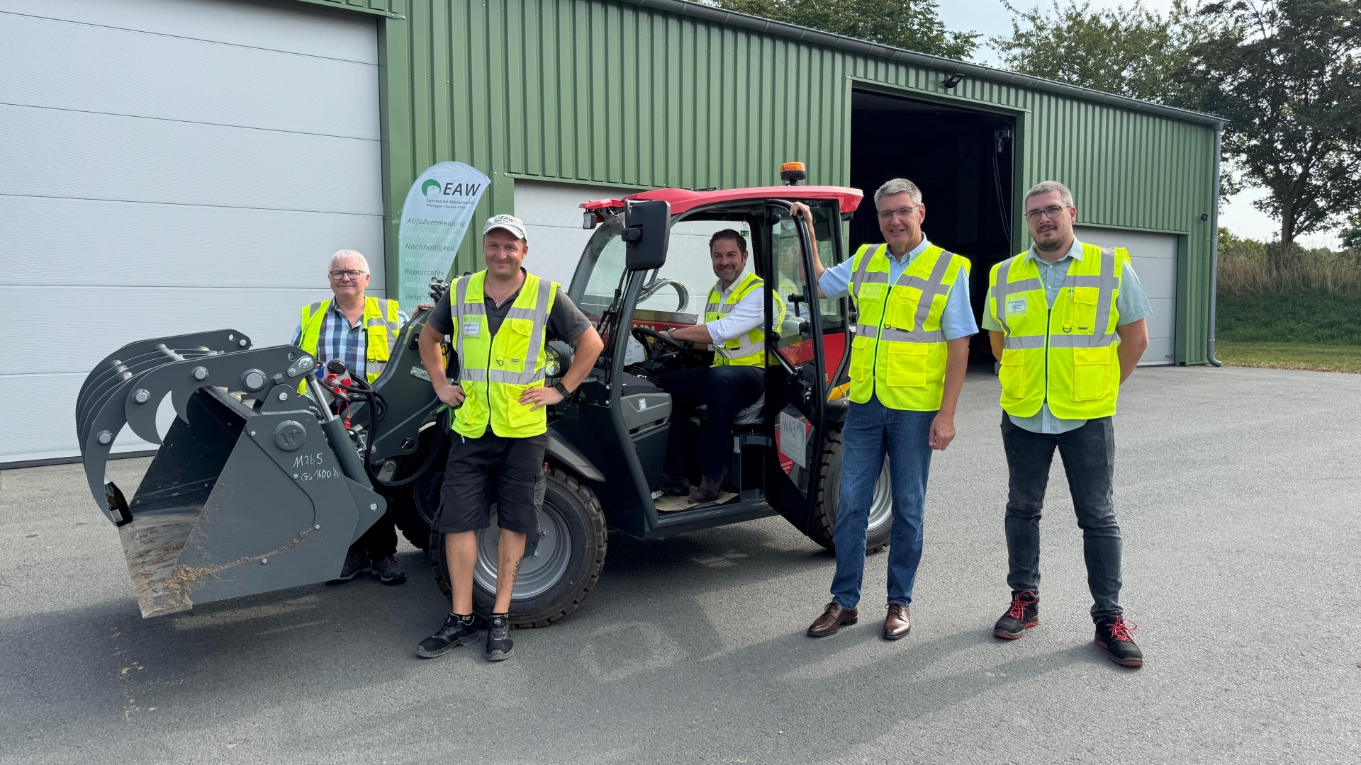 Vor einem großen, grünem Gebäude mit vier großen geschlossenen Toren und einem offenen Tor stehen vier Männer mit neongelben Warnwesten vor und hinter einem Radlader mit Elektroantrieb. Auf dem Sitz des Radlader sitzt ein weiterer Mann mit neongelber Warnweste. Der Radlader hat ein rotes Dach. Im Hintergrund sind Bäume und Wiese und vor dem Gebäude steht eine Beachflag.