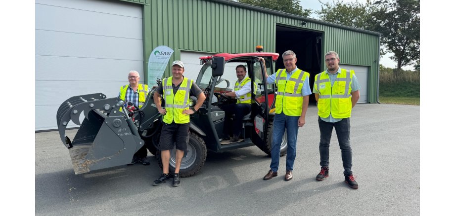 Vor einem großen, grünem Gebäude mit vier großen geschlossenen Toren und einem offenen Tor stehen vier Männer mit neongelben Warnwesten vor und hinter einem Radlader mit Elektroantrieb. Auf dem Sitz des Radlader sitzt ein weiterer Mann mit neongelber Warnweste. Der Radlader hat ein rotes Dach. Im Hintergrund sind Bäume und Wiese und vor dem Gebäude steht eine Beachflag.