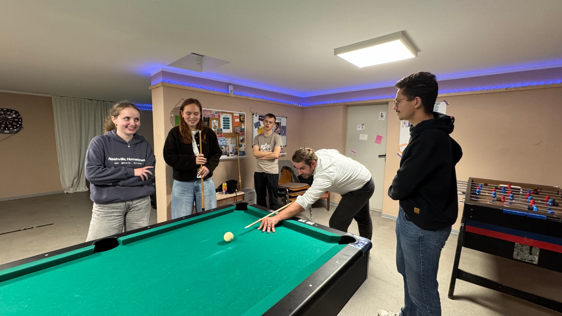 In einem großem Raum hängt eine Dartscheibe und eine Pinwand an der Wand und es steht ein Bililardtisch und ein Tischkicker im Raum. Um den Tisch stehen vier Jugendliche und ein Mann, der den Kö in der Hand hält und kurz davor ist, mit dem Stoß zur weißen Kugel, das Spiel zu beginnen. 