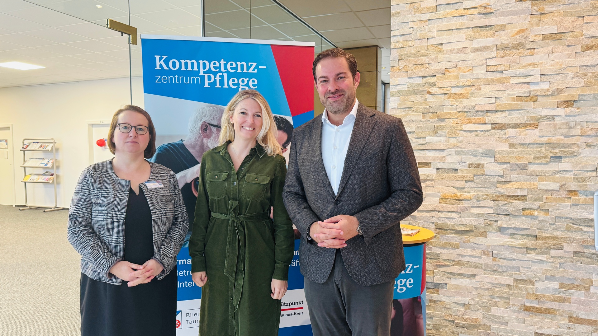 Vor einem Plaket mit der Aufschrift "Kompetenzzentrum Pflege stehen zwei Frauen und ein Mann. Die Frau in der Mitte hat blonde, lange Haare und ein grünes langes Kleid und die Frau daneben braune kinnlange Haare und eine Brille und ein schwarzes Kleid mit einer schwarz-weiß karrierten Jacke. Der MAnn trägt ein weißes Hemd und eine sunkelgraue Hose und Jacket. Rechts ist eine Steinwand zu sehen und links ein Flur mit einem Plakatständer.    