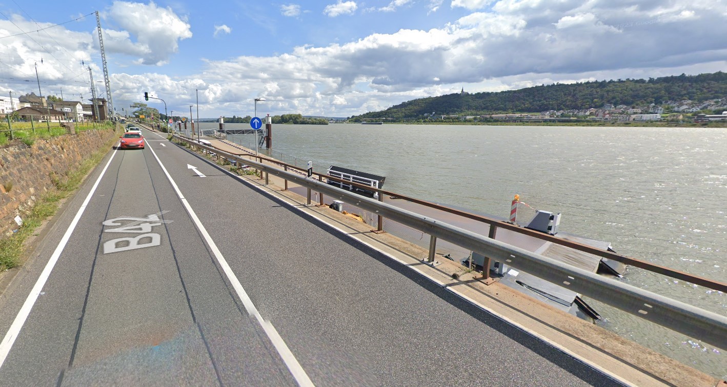 Man sieht die Bundesstraße 42, mit der Bahnstrecke linkerhand. Rechts davon ist der Rhein und die andere Rheinseite mit Gebäuden und viel Wald. Neben der Straße ist der Fähranleger zu sehen. Auf der Straße fahren Autos und man sieht eine grüne Ampel und Verkehrsschilder. Der Himmel ist blau mit weißen Wolken.