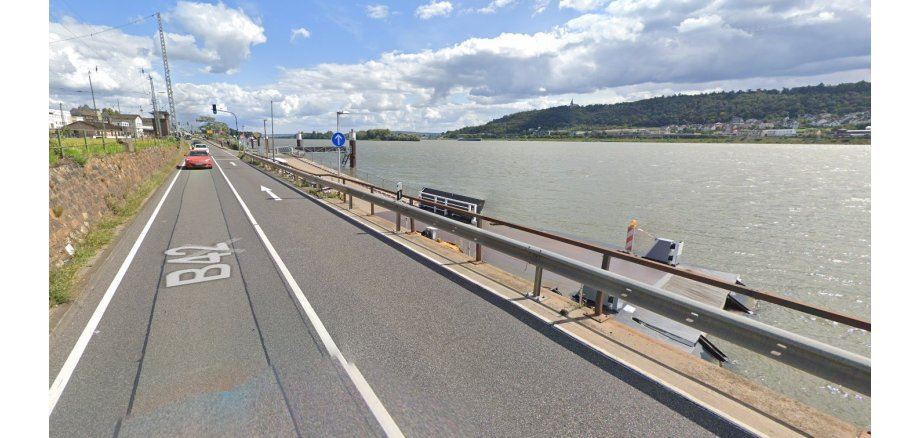 Man sieht die Bundesstraße 42, mit der Bahnstrecke linkerhand. Rechts davon ist der Rhein und die andere Rheinseite mit Gebäuden und viel Wald. Neben der Straße ist der Fähranleger zu sehen. Auf der Straße fahren Autos und man sieht eine grüne Ampel und Verkehrsschilder. Der Himmel ist blau mit weißen Wolken.