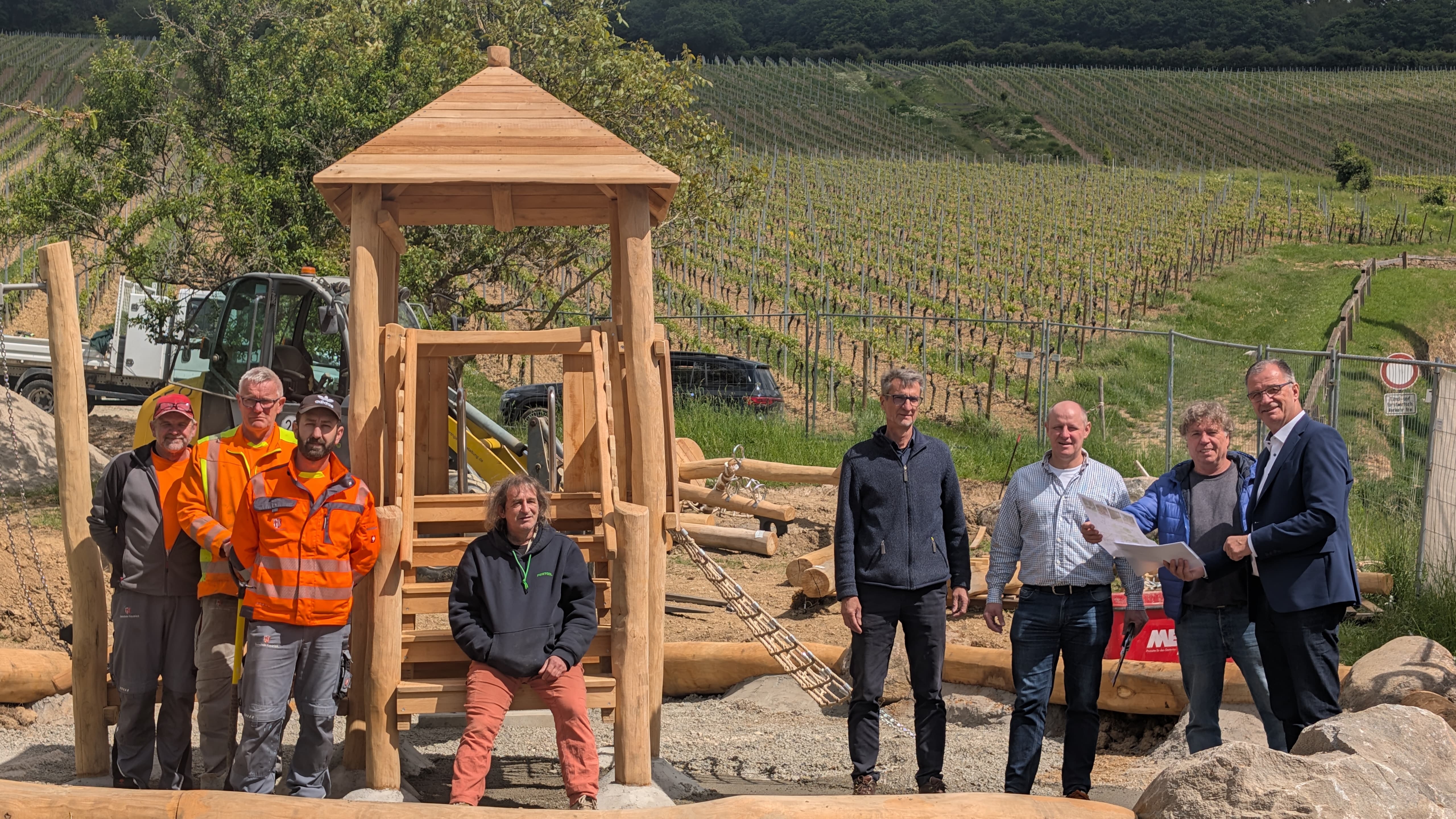Acht Herren, darunter Bürgermeister Steinmacher, stehen vor einem Holzspielturm, mit einem Kletternetz an der Seite. Drum herum liegt weiteres Holz und Steine und Kies. Die Baustelle ist von Bauzäunen umschlossen. Im Hintergrund sind Weinberge.