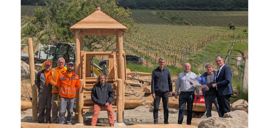 Acht Herren, darunter Bürgermeister Steinmacher, stehen vor einem Holzspielturm, mit einem Kletternetz an der Seite. Drum herum liegt weiteres Holz und Steine und Kies. Die Baustelle ist von Bauzäunen umschlossen. Im Hintergrund sind Weinberge.