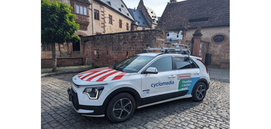 Ein weißes Fahrzeug mit Folienwerbung und zwei Kameras auf dem Dachgepäckträger steht auf Kopfsteinpflaster vor einem Laubbaum, historischen Gebäuden und einer alten Steinmauer mit Toren aus Eisenstäben und Wappentieren aus Stein auf den Eingangssäulen.