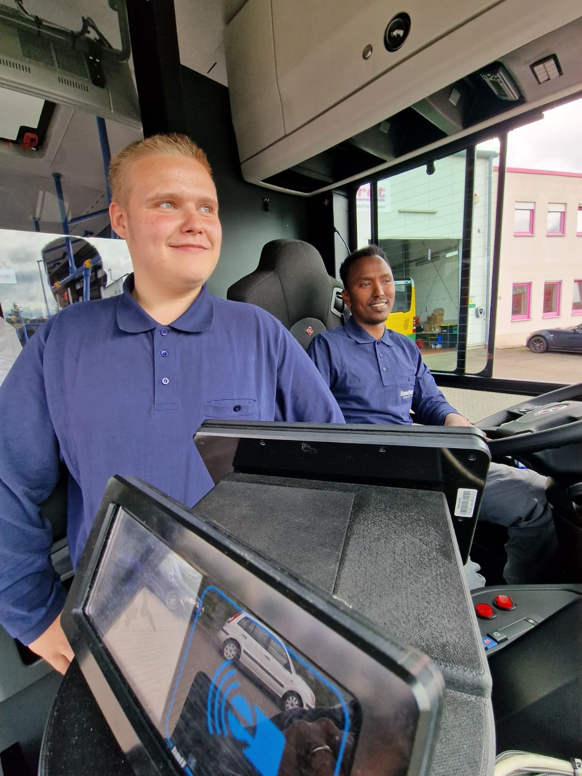 In einem Linienbus sitzen zwei junge Männer in blauem, langärmligem Polo-Shirt. Der Eine sitzt hinter dem Lenkrad und der andere daneben. Im Vordergrund ist das Display der Fahrzeugkamera mit einem Auto zu sehen.