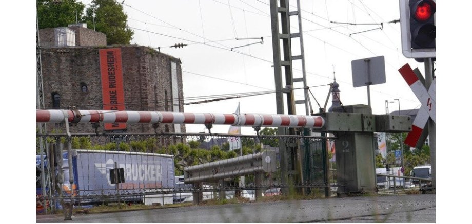 Ein Bahnübergang mit geschlossener Schranke, roter Ampel und Andreaskreuz. Im Hintergrund steht ein blauer LKW und eine Burg mit einem langem roten Banner und ein Kirchturm. Die Stromleitungen der Bahn und ein Mast sind oberhalb der Schranke.