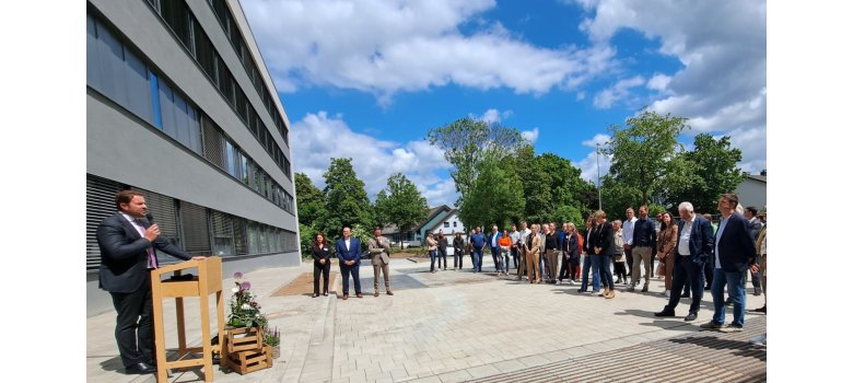 Landrat Sandro Zehner (l.) bei der Einweihung des neuen Gebäudes der Beruflichen Schulen Rheingau. Vor einem großem grauen Schulgebäude stehen auf einer gepflasterten Fläche mehrere Menschen, die einem Mann im Anzug am Rednerpult zuhören. Vor dem Rednerpult stehen zwei Kisten, auf denen ein hohes Blumengesteck steht.