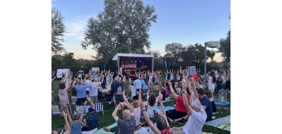 Auf einer Wiese, auf der auch Bäume stehen, ist eine Bühne aufgebaut, auf der eine Band spielt. Auf der Wiese stehen und sitzen Menschen auf Picknickdecken und Campingstühlen. Alle Leute haben die Arme nach oben gestreckt.