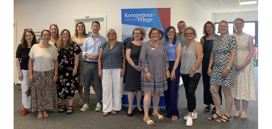 Gruppenbild Jahrestreffen In einem Flur mit Türen und Fenstern stehen 2 Männer und 13 Frauen vor einem Roll-up in sommerlicher Kleidung.