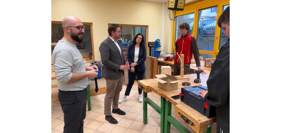 In einem Raum mit Fenstern mit gelben Fensterrahmen, stehen mehrere Werkbänke mit Holzplatten und grünen Metallbeinen. Davor stehen vier Männer und eine Frau.