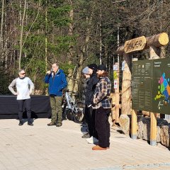 Man sieht Bürgermeister Steinmacher, wie er in ein Mikrofon spricht. Daneben stehen vier Männer und ein Mountainbike vor dem Eingang zur Mountainbike-Strecke, der aus großen Holzstämmen zusammen gebaut wurde. Oben hängt ein Holzschild mit "Trailhead". Rechts davon ist eine Hinweistafel u.a. mit den Routen und Öffnungszeiten an drei großen Holzpfählen angebracht. Im Hintergrund ist der Wald.