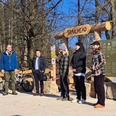Man sieht Bürgermeister Steinmacher, sowie fünf Männer und ein Mountainbike vor dem Eingang zur Mountainbike-Strecke, der aus großen Holzstämmen zusammen gebaut wurde. Oben hängt ein Holzschild mit "Trailhead". Rechts davon ist eine Hinweistafel u.a. mit den Routen und Öffnungszeiten an drei großen Holzpfählen angebracht. Im Hintergrund ist der Wald.