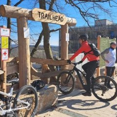 Man sieht den Eingang zur Mountainbike-Strecke, der aus großen Holzstämmen zusammen gebaut ist. Oben hängt ein Holzschild mit "Trailhead". Es sind vier Mountainbikes und zwei Fahrer zu erkennen im Hintergrund unterhalten sich zwei Männer. Ganz hinten ist die Fassade des WALD-WEIT Hotel zu erkennen.