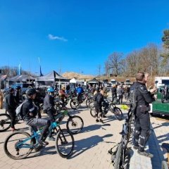 Auf einem asphaltierten Parkplatz stehen mehrere Personen und viele Mountainbikes. Es sind mehrere Pavillons und mehrere Dropflags aufgestellt. Im Hintergrund sind Bäume und ein blauer Himmel.