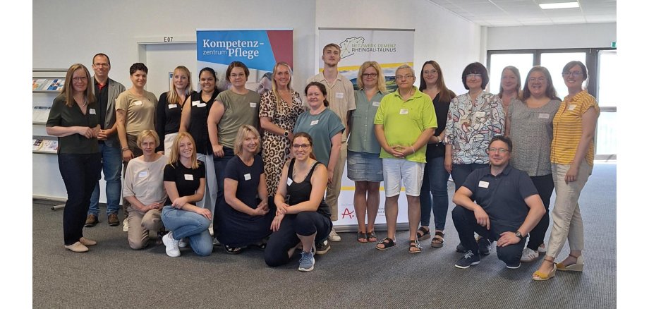 Eine Gruppe, bestehend aus 21 Personen, stehen in einem Flur mit einem Prospektständer und zwei RollUp-Banner. Jeder trägt ein weißes Namensschild.