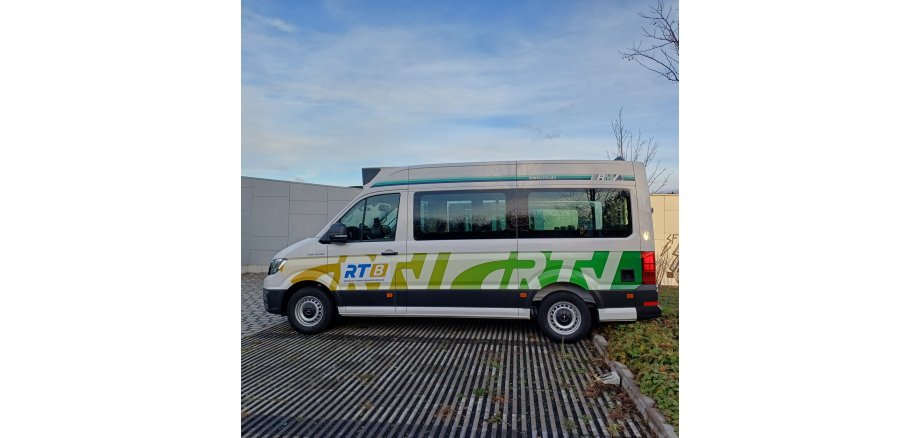 Auf einem Parkplatz mit Betonrillen steht ein weißer Kleinbus mit dem hell- und dunkel-grünem RTV-Logo. Der Himmel ist blau mit weißen Wolgen. An den Bäumen sind keine Blätter mehr.