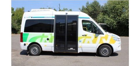 Auf einer Asphaltfläche, vor grünen Laubbäumen und Hecken, steht ein Kleinbus in weiß und großen schwarzen Schiebetüren. Das Logo RTV ist ebenso weiss, aber grün/gelb umrandet.