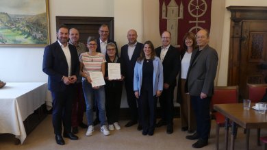 In einem Raum mit zwei alten braunen Türen hängt an der Wand eine Stoffbahn mit einem Wappen in rot, mit beigem Turm und Doppelrad auf beigem Untergrund und ein altes Landschaftsgemälde mit goldenem Rahmen. Davor stehen vier Frauen und 6 Männer überwiegend in dunkeln Anzügen und weißen Hemden.