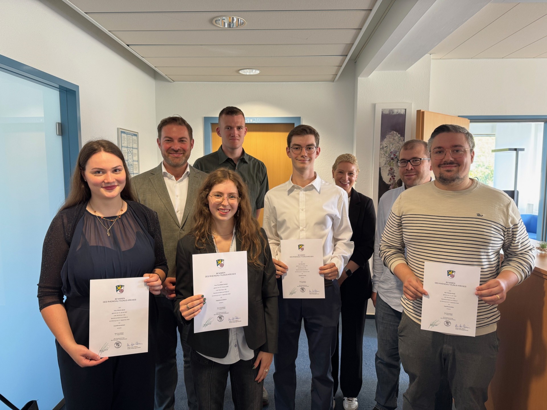 Zwei junge Frauen und zwei junge Männer halten weiße Urkunden in den Händen. Dahinter steht eine Frau und drei Männer. Im Hintergrund links ist eine geschlossene Glastür, geradeaus eine geschlossene Holztür und rechts davon eine geöffnete Holz-Büro-Tür. Die Türrahmen sind blau umrandet und die Wände sind weiß.
