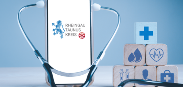 Auf einem Handy-Display erscheint das Rheingau-Taunus-Logo. Um das Handy ist ein Stethoskop. Daneben sind Holzwürfel, drei unten, zwei darüber und oben ein Würfel. Jeder Würfel hat ein blaues Zeichen, ein Kreuz, zwei Tropfen, ein Arztkoffer, ein Herz mit einer Herzlinie, ein Mann und zwei Spritzen.