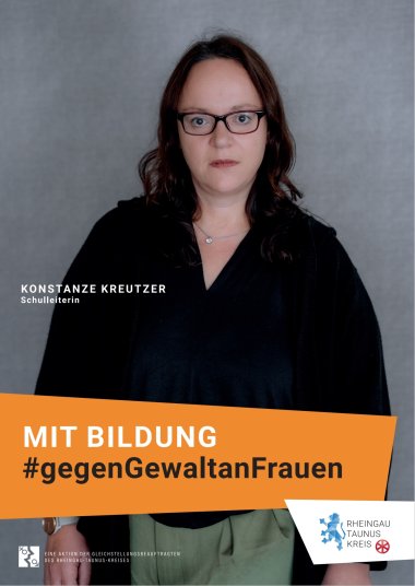 Vor einem grauen Hintergrund steht eine Frau mit schwarzer Bluse und oliv grüner Hose. Sie trägt eine silberne Kette mit einem Glitzerstein. Die braunen Haare sind schulterlang und offen. Sie trägt eine dunkelbraune, rechteckige Brille. Der Blick ist ernst.