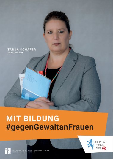 Vor einem grauen Hintergrund steht eine Frau im grauem Blazer, die zwei Schulbücher mit verschränkten Armen vor ihren Körper hält. Sie trägt eine schwarze Hose und ein schwarzes Shirt mit einer bunten Perlenkette. Die braunen Haare sind zu einen Zopf zurückgebunden. Der Blick ist ernst.