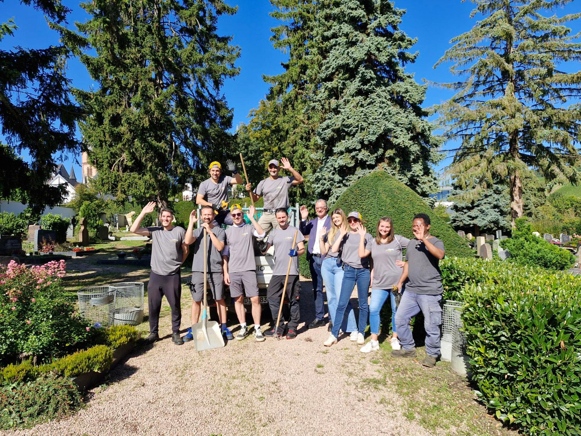Auf einem Weg mit kleinen Splittsteinen, stehen 11 Personen. Ein Mann in Jeans und weißem Hemd und blauem Jacket. Die anderen mit grauen T-Shirts mit dem Logo der Süwag und Arbeitshosen. Alle winken und einige haben einen Spaten oder eine Hacke in der Hand. Im HIntergrund sind Hecken, Gräber, Kirschlorbeerbüsche und große Nadelbäume zu sehen. Der Himmel ist blau und ohne Wolken.