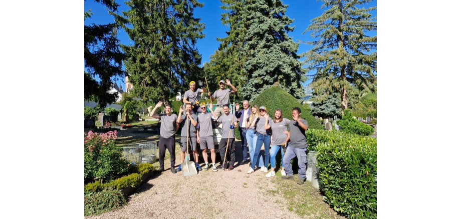 Auf einem Weg mit kleinen Splittsteinen, stehen 11 Personen. Ein Mann in Jeans und weißem Hemd und blauem Jacket. Die anderen mit grauen T-Shirts mit dem Logo der Süwag und Arbeitshosen. Alle winken und einige haben einen Spaten oder eine Hacke in der Hand. Im HIntergrund sind Hecken, Gräber, Kirschlorbeerbüsche und große Nadelbäume zu sehen. Der Himmel ist blau und ohne Wolken.