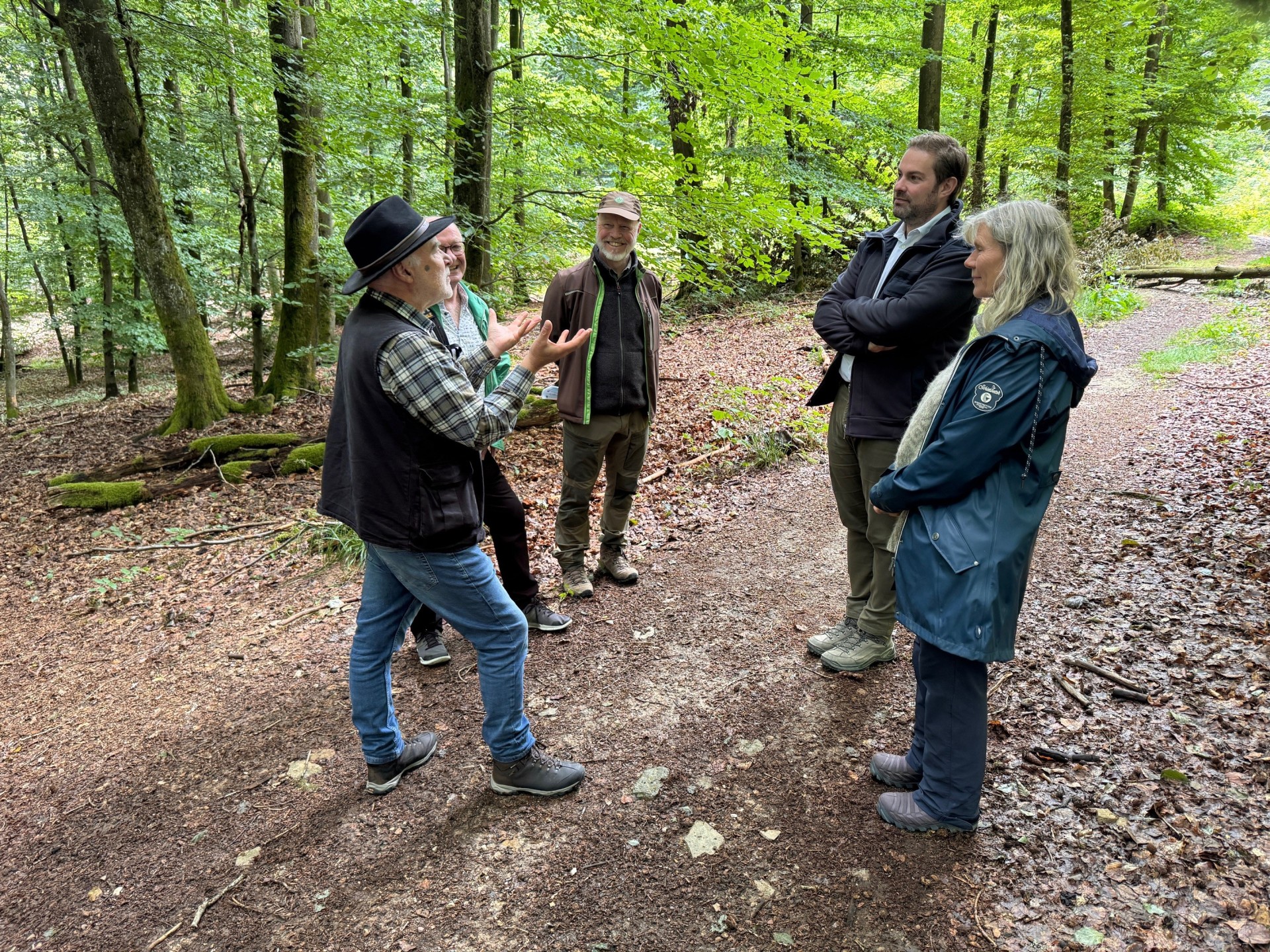 Auf einem Waldweg, auf dem am Rande Laubbäume sind, stehen vier Männer und eine Frau im Kreis und unterhalten sich miteinander. Der Waldboden und der Weg ist mit braunen, alten Blättern bedeckt. 