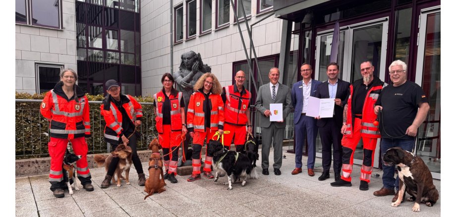 Vor einem Verwaltungsgebäude mit einem großem Glastreppenhaus und vielen Fenstern, stehen 6 Männer und vier Frauen mit sechs Hunden. Drei der Männer haben Anzüge und Hemden an und halten Urkunden in den Händen, der Rest hat orangene Hosen und Jacken mit Reflektierstreifen an. Ganz außen der Mann trägt ein schwarzes T-Shirt und eine blaue Jeans. Im Hintergrund ist eine Statue aus Stein zu erkennen.