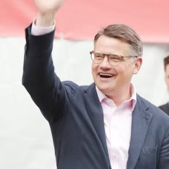 Ein Mann mit rosa-weiß gestreiftem Hemd und dunklem Anzug winkt und lacht. Er hat kurze Haare und eine Brille.