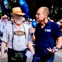 Ein Mann mit kurzen Haaren und blauem Polo-Shirt und Mikrofon interviewt einen Mann im weißem Hemd, Lederhose und Sonnenhut mit rotem Band.  