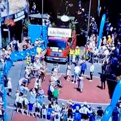 Blick von oben auf ein Blasorchester mit Instrumenten und dem rotem LKW mit großem weißem Schild auf dem Führerhaus und Puplikum hinter den Absperrungen.