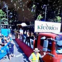 Ein roter Laster mit Holzbalken, runden Schildern und Blumen geschmückt fährt auf einer Allee. Neben dran sind Menschen hinter den Absperrungen. Am Seitenspiegel läuft ein Mann mit Sonnenhut und Warnweste.