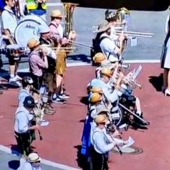 Eine Fußgruppe läuft mit Lederhosen und Hemden und Sonnenhüten über einen großen roten Kreis. Die Menschen der Musikgruppe  haben Musikinstrumente in der Hand und bespielen sie. 