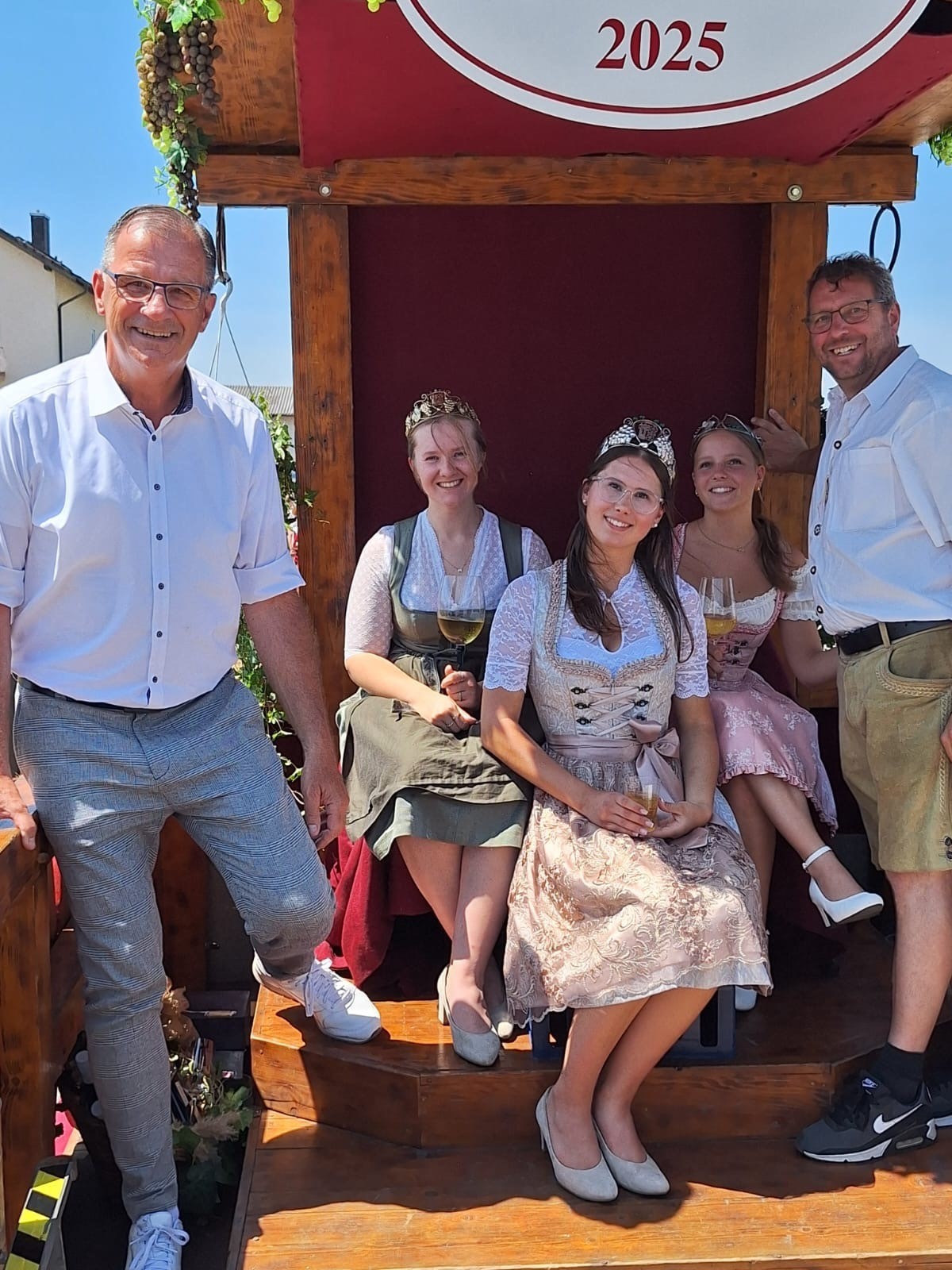 EIn Mann mit kurzärmeligem, weißem Hemd und grau, weiß karrierten Hose und ein weiterer Mann ebenso in weißem Hemd und kurzer Hose, stehen links und rechts von drei jungen Frauen mit Kronen auf dem Kopf und Weinglas in der Hand. Sie tragen Dirndl und weiße Dirndlblusen.