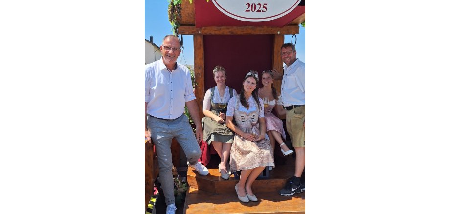 EIn Mann mit kurzärmeligem, weißem Hemd und grau, weiß karrierten Hose und ein weiterer Mann ebenso in weißem Hemd und kurzer Hose, stehen links und rechts von drei jungen Frauen mit Kronen auf dem Kopf und Weinglas in der Hand. Sie tragen Dirndl und weiße Dirndlblusen.