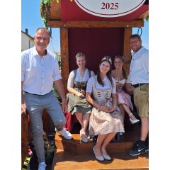 EIn Mann mit kurzärmeligem, weißem Hemd und grau, weiß karrierten Hose und ein weiterer Mann ebenso in weißem Hemd und kurzer Hose, stehen links und rechts von drei jungen Frauen mit Kronen auf dem Kopf und Weinglas in der Hand. Sie tragen Dirndl und weiße Dirndlblusen.