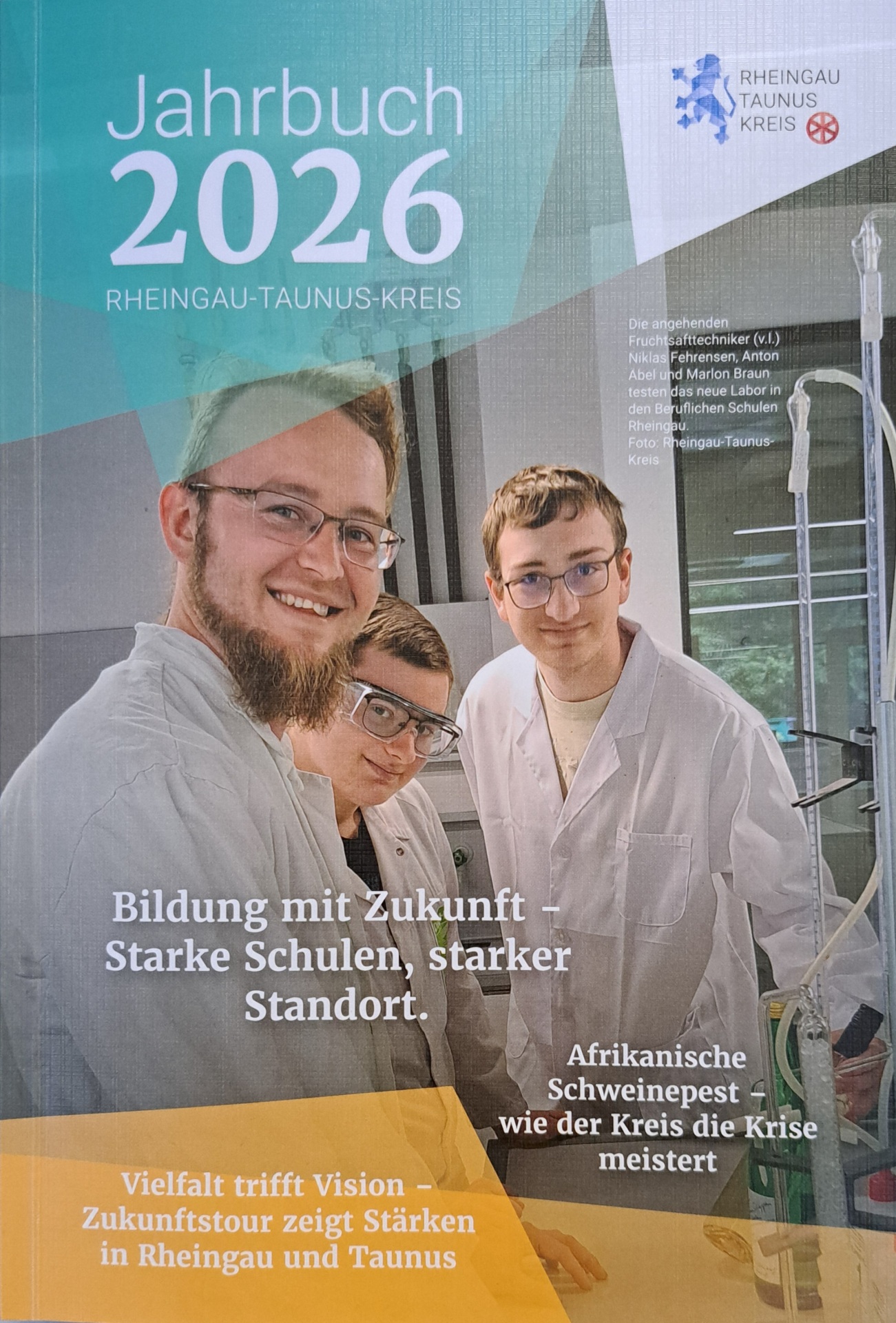 Titelblatt des Jahrbuches 2026 des Rheingau-Taunus-Kreises auf dem drei junge Männer in weißen Kitteln mit kurzen blonden Haaren mit zwei normalen Brillen und einer Schutzbrille in einem Labor stehen. Ein Mann trägt Bart.