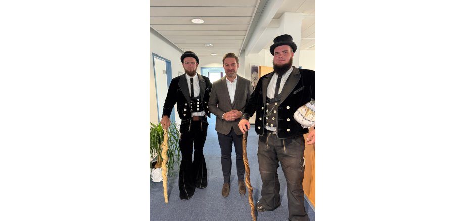 Landrad Zehner steht im Anzug zwischen zwei Gesellen auf der Walz. Sie tragen den schwarzen Hut, die Traditionskleidung in schwarz mit weißen Knöpfen, die Weste und das weiße Hemd. Beide tragen einen langen Bart und haben geschwungene Wanderstöcke inn der Hand.