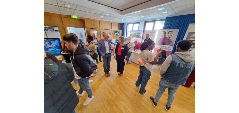 In einem Raum stehen mehrere Menschen und sprechen miteinander. Es sind InfoTische mit Flyern aufgestellt, hinter denen Roll-Ups mit Werbung für Firmen für einen Ausbildungs- oder Arbeitsplatz stehen.