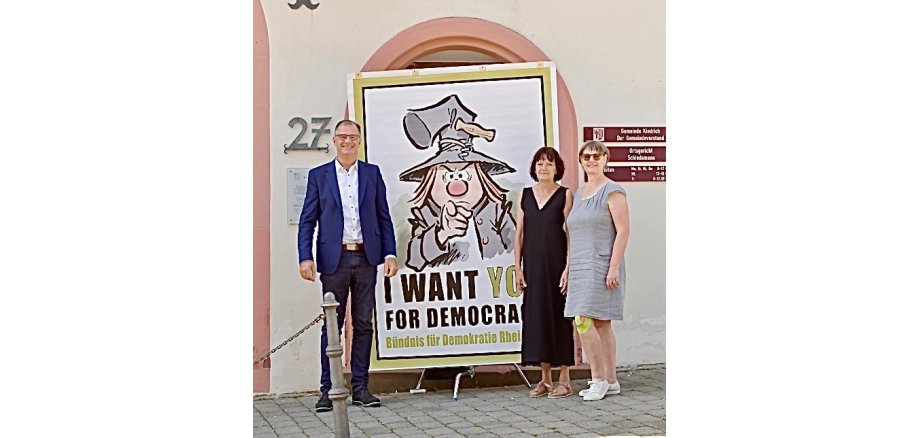 Bürgermeister Steinmacher und zwei Frauen in Kleidern stehen vor dem Rathaus und vor einem Plakat, auf dem ein gezeichneter Mann mit grauem Schlappgut und einem Korkenzieher an der Hutkrempe und einer grauen Jacke abgemildet ist. Er zeigt mit dem Finger auf den Betrachter, schaut grimmig und unter ihm steht "I WANT YOU FOR DEMOCRATY".