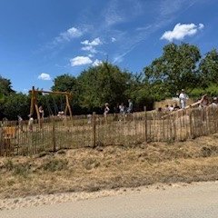 Im Vordergrund ist eine Straße und vertrocknete Wiese. Dahinter einen Holzzaun und dahinter ein Spielplatz mit mehreren Mensche. Es ist eine Holzwippe und eine Mehrgenerationsschaukel zu erkennen. Dahinter sind Bäume und Weinberge. Der Himmel ist bewölkt und hat wenige weiße Wolken.